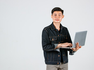 Portrait studio shot of Asian young handsome male hipster model wearing casual street denim jeans jacket standing holding laptop notebook computer typing surfing browsing internet on white background