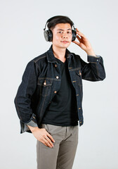 Portrait studio shot of Asian happy handsome male hipster model wearing casual street denim jeans jacket and headphone standing holding smartphone listening to music songs playlist on gray background