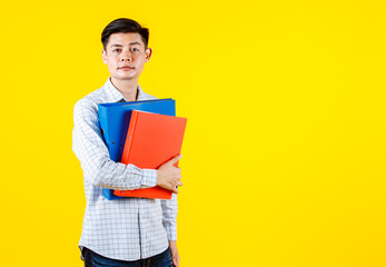 Portrait studio shot of Asian young male businessman employee model in casual outfit standing look at camera holding paperwork document file colorful hard case folders in arm on yellow background