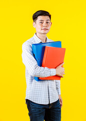Portrait studio shot of Asian young male businessman employee model in casual outfit standing look at camera holding paperwork document file colorful hard case folders in arm on yellow background