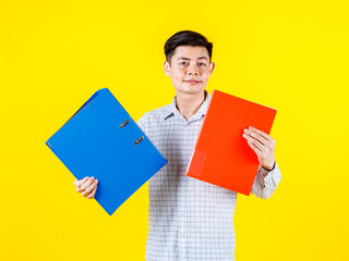 Portrait studio shot of Asian young male businessman employee model in casual outfit standing look at camera holding paperwork document file colorful hard case folders in arm on yellow background