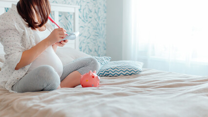 Pregnant woman writing down in notebook on bed. Woman making shopping list. Pregnant woman counting money from piggy bank.