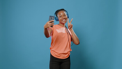 Naklejka premium Modern woman recording dance moves and singing song on headphones in studio. Young adult smiling and holding smartphone to film video on camera. Person using headset and technology.