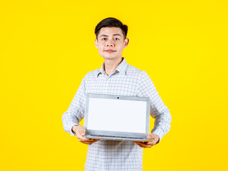 Portrait studio shot of millennial Asian young male businessman in casual outfit standing smiling showing white blank empty screen laptop computer for advertising copy space on yellow background