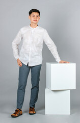 Portrait studio shot millennial Asian happy handsome male model in casual long sleeve shirt outfit and leather sneakers standing posing on white square stool smiling look at camera on gray background