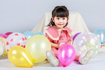 Fototapeta na wymiar Portrait studio shot of little cute kindergarten preschooler kid girl daughter in red long dress sitting smiling alone on floor playing with colorful helium air party balloons on gray background
