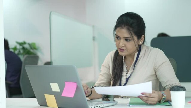 Young serious Indian ethnicity woman working on laptop reading reports on papers,comparing information,check list of tasks,do paperwork,studying or telecommuting from office using modern tech concept.
