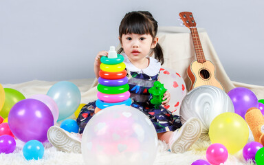 Studio shot of little cute innocent kindergarten preschooler girl daughter in casual dress sitting between colorful helium balloons playing dinosaur and plastic rings pyramid toys on gray background