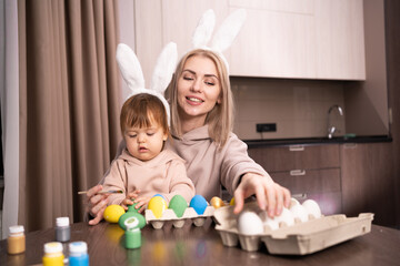 Happy mother and daughter are painting Easter eggs while sitting at the table. Easter craft. woman and girl paint easter eggs. Happy family is preparing for Easter. authentic portrait.