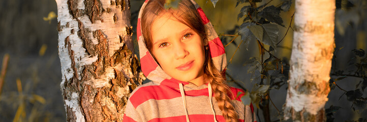 portrait of candid eight year old kid girl in striped gray red hoodie near the trunk of  birch tree in summer nature on a hike in the warm rays of the setting sun at the golden hour at sunset. banner