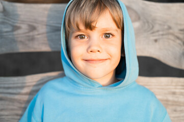 portrait of face happy smiling candid five year old kid boy in blue hoodie in a hood on the background of wooden boards in the nature of summer in a golden hour at sunset. homeschool and travel