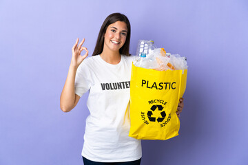 Young brazilian woman holding a bag full of plastic bottles to recycle isolated on purple background showing ok sign with fingers