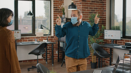 Fototapeta na wymiar Happy employee dancing after winning successful deal, wearing face mask. Business man celebrating achievement and feeling excited while woman looking at playful colleague. Funny worker