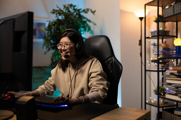 Smiling girl in sweatshirt and glasses with headphones plays video games online with friends talking to them through microphone, pass levels, rounds, strategy, victory, gamer room led neon lighting