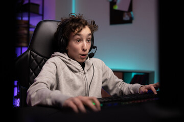 Surprised school-age gamer with headset boy sits late at night in front of computer monitor playing multiplayer shooter video games with team excitement of virtual world room lit up with led lights © ABCreative