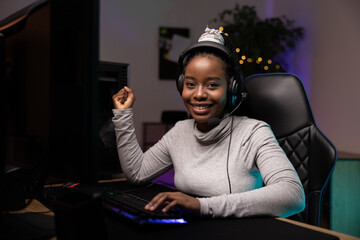 Pro gamer girl with dark skin plays a pc game with a team she talks to through a headset, the team wins a round in an online virtual championship, the girl clenches fists in joy at the victory