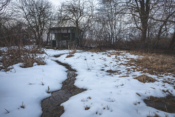 small ditch with water leads to abandoned wooden garden house. Snow covered ground in early spring - Powered by Adobe