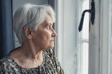 grey-haired sad upset elderly woman in her 60s looking out of the window medium close up indoor...