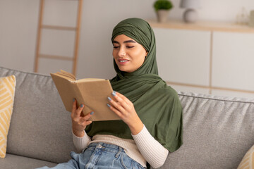 Peaceful domestic hobbies. Cheery young Arab woman in hijab reading book, relaxing on sofa in living room