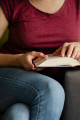 Fototapeta premium Selective focus on book read by caucasian woman in burgundy T-shirt and jeans sitting on gray sofa with pillows. Concept of rest in cozy house. Time alone with yourself.