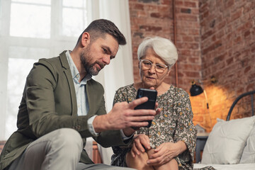 middle-aged caucasian son learning his pensioner mother how to use a smartphone . High quality photo