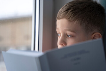 A cute Caucasian boy of 5 years old in a red T-shirt sits on the windowsill by the window with a book