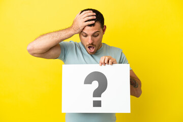 Brazilian man over isolated purple background holding a placard with question mark symbol with surprised expression