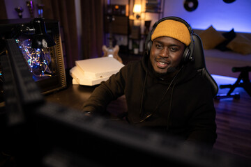 Men's room at night lit by led lights. A pro gamer sits in front of a computer playing online internet games with a team he's talking to through a headset. A relaxed guy addicted to gaming.