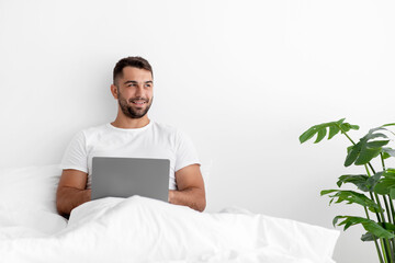 Cheerful millennial caucasian handsome male sitting on white bed, thinking, creating idea and typing on computer