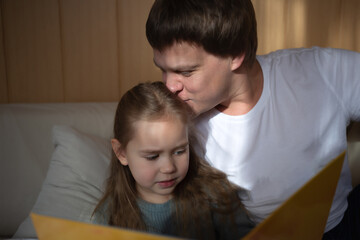 little cute girl with dad read a book sitting in the bedroom on the couch