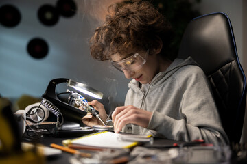 Little boy with safety glasses, future electronics technician, learning how to solder microprocessor cables and printed circuits, looking through a magnifying glass, repairing and testing electronics