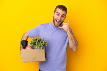 Young caucasian making a move while picking up a box full of things isolated on yellow background making phone gesture. Call me back sign