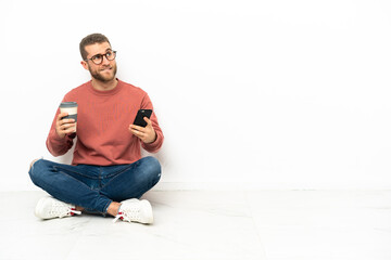 Young handsome man sitting on the floor holding coffee to take away and a mobile while thinking something