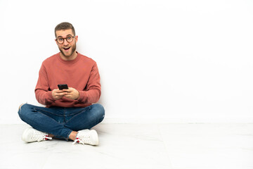 Fototapeta premium Young handsome man sitting on the floor surprised and sending a message