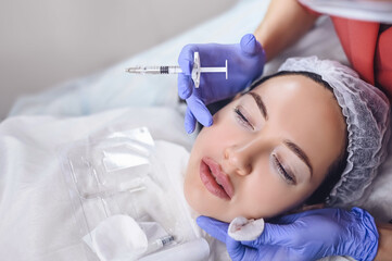 Fototapeta premium Cosmetologist in beauty salon doing lip augmentation procedure with hyaluronic acid. The beautician pierces the lips with a needle. Young woman doing subcutaneous injection to increase lips shape.