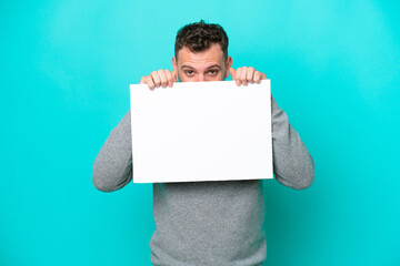 Young Brazilian man holding an empty placard isolated on blue background holding an empty placard and hiding behind it