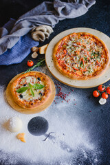 Photo of pizza on a textured wooden table in a restaurant. There are various ingredients nearby: cheese, mushrooms, tomatoes, spices, flour.
