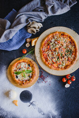 Photo of pizza on a textured wooden table in a restaurant. Top view. There are ingredients nearby: cheese, mushrooms, tomatoes, spices, flour.