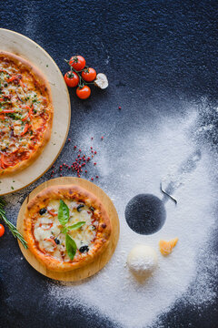 Photo of pizza on a textured wooden table in a restaurant. Top view. There are various ingredients nearby: cheese, mushrooms, tomatoes, spices, flour