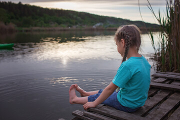 Fototapeta premium Happy little girl sitting on pontoon bridge on the bank of lake and enjoying warm sunny evening, childhood in village in summer, countryside