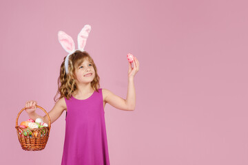 Adorable little girl with blond curly hair in stylish dress and bunny ears, smiling and looking away while demonstrating wicker basket and colorful Easter egg