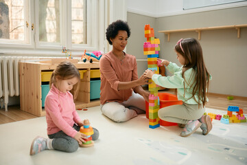 Happy black teacher and preschool kids play with toy blocks in playroom.