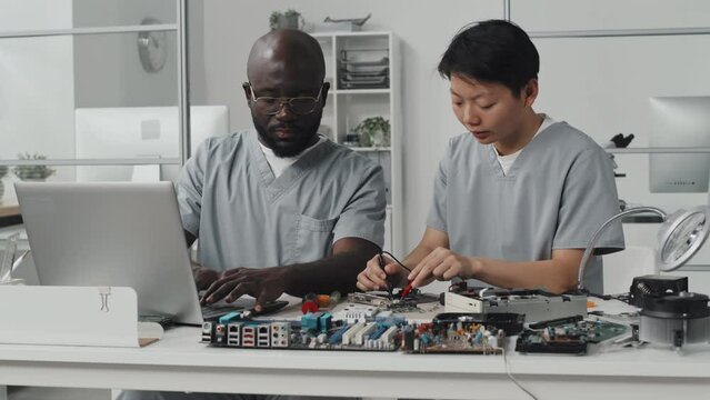 Asian female engineer using multimeter and talking with African American colleague as he working on laptop at desk in laboratory