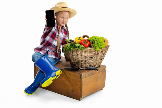 Beautiful cute little girl, emotive kid in image of farmer, gardener with large basket of vegetables using phone isolated on white background.