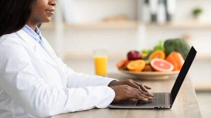 Black female dietitian with laptop consulting patient online at clinic, sitting at desk, side view,...