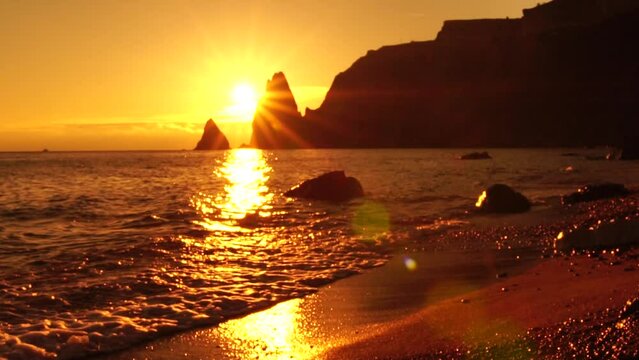 A red burning sunset over the sea with rocky volcanic cliff. Abstract nature summer or spring ocean sea background. Small waves on golden warm water surface with bokeh lights from sun.