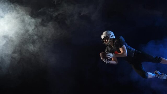 American football player in uniform, pads and helmet, catching pass and diving to ground with ball. Determination quarterback plays American football in dark arena among smoke and lights. Slow motion.