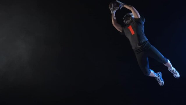 American football player in uniform, pads and helmet, jumping, catching pass and runs with ball across field. Quarterback plays American football in dark arena among smoke and lights. Slow motion.