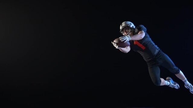 American football player wearing uniform, pads and helmet, jumping, catching pass and diving to ground with ball. Determination quarterback plays American football on a black background. Slow motion.