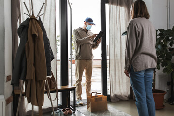 Fototapeta premium food shipping, pandemic and people concept - delivery man in mask with tablet pc computer and paper bag and female customer at home